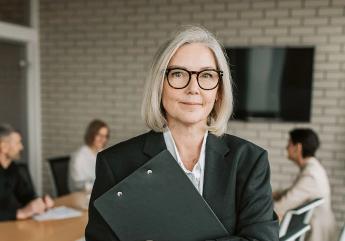 Portrait professionnel d'une femme cadre d'âge mûr avec des lunettes, tenant une chemise de documents, avec son équipe travaillant dans une salle de réunion moderne en arrière-plan. Idéal pour les sites d'entreprise, le consulting et le management.