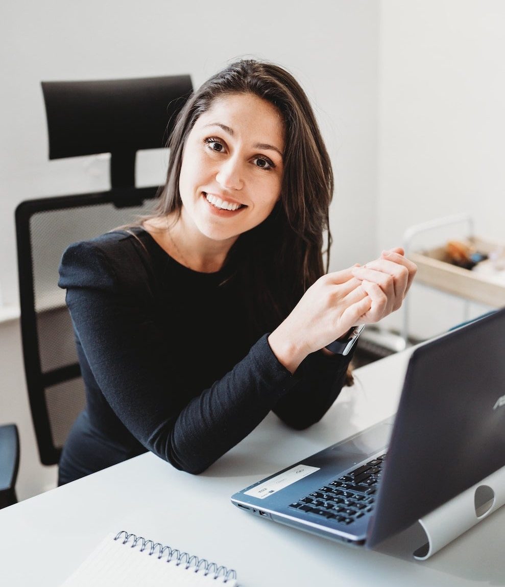 Photo de profil professionnelle d'une femme souriante, les mains croisées sur un bureau à côté d'un ordinateur portable, présentée dans un format circulaire. Idéal pour illustrer l'équipe, le contact ou l'accueil client.