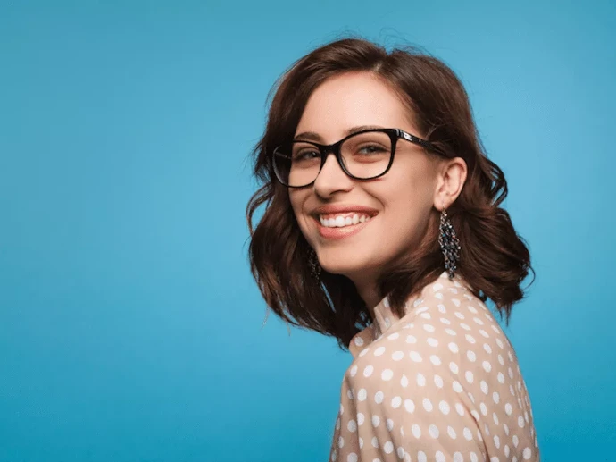 Photographie de portrait lumineuse et chaleureuse d'une femme souriante aux cheveux bruns bouclés, portant une blouse à pois et des lunettes de vue, sur un fond uni bleu vif. Parfait pour illustrer la relation client, l'accueil ou un service de proximité.