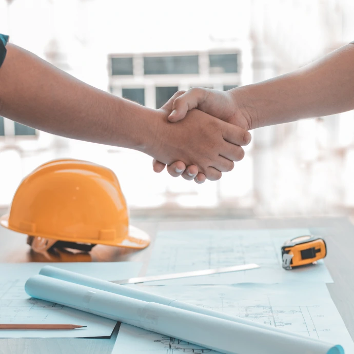 Deux personnes se serrant la main pour sceller un accord professionnel, posées sur une table de réunion jonchée de plans architecturaux et d'un casque de chantier jaune. Idéal pour illustrer les partenariats B2B, la conclusion de contrats et la collaboration dans le secteur technique.